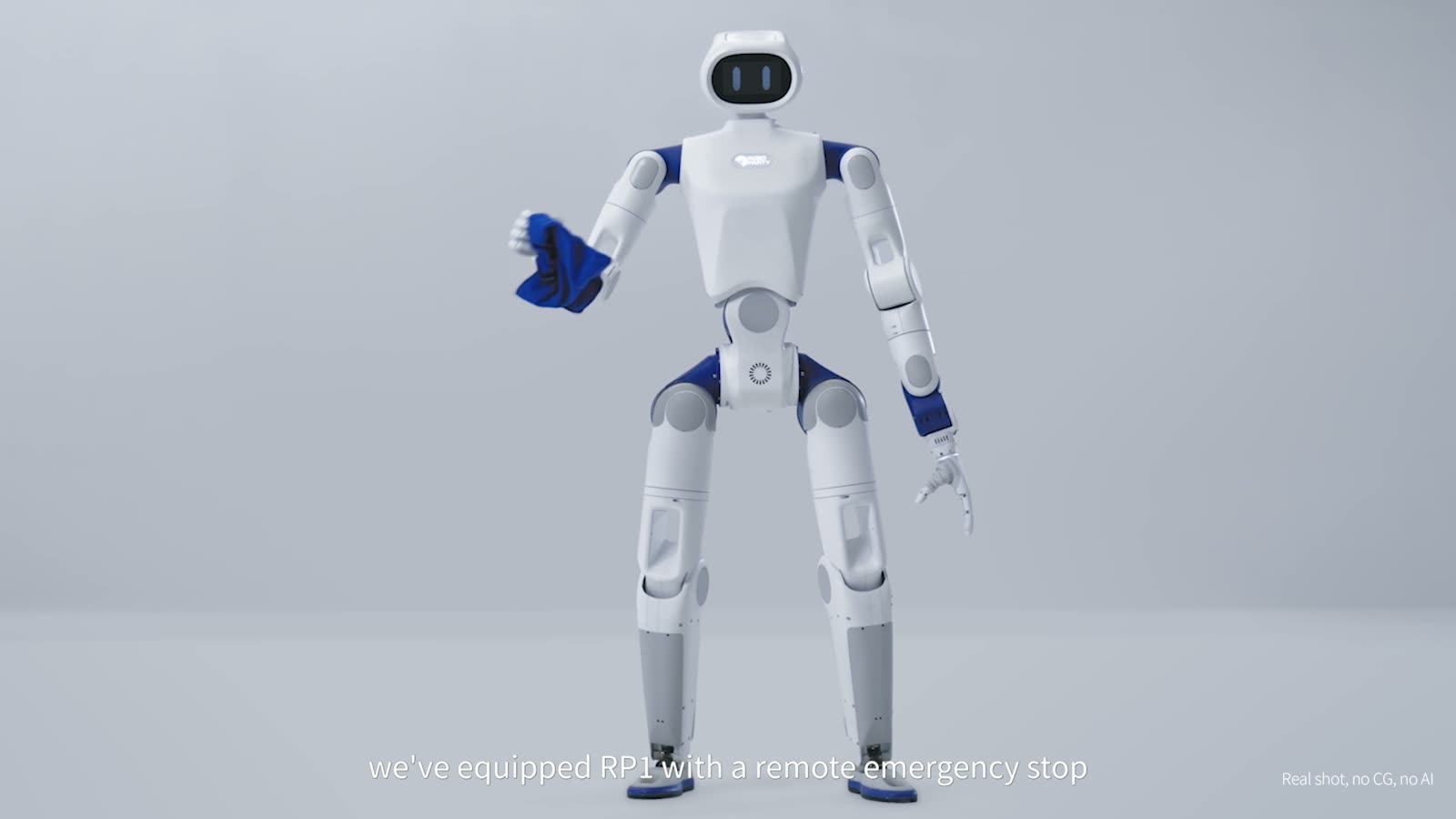 The white and blue RoboParty RP1 humanoid robot standing in a plain grey studio, holding a blue cloth in one hand