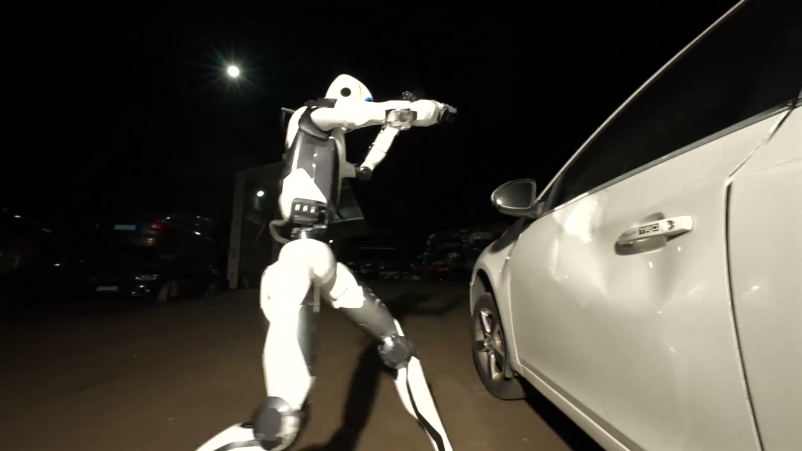 The white Unitree H2‑X humanoid robot kicking the door of a white car at night